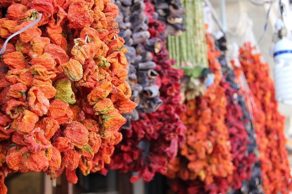 Close-up of colorful strings of dried vegetables, including bright orange, red, and deep purple peppers, hanging in bunches at a market stall.