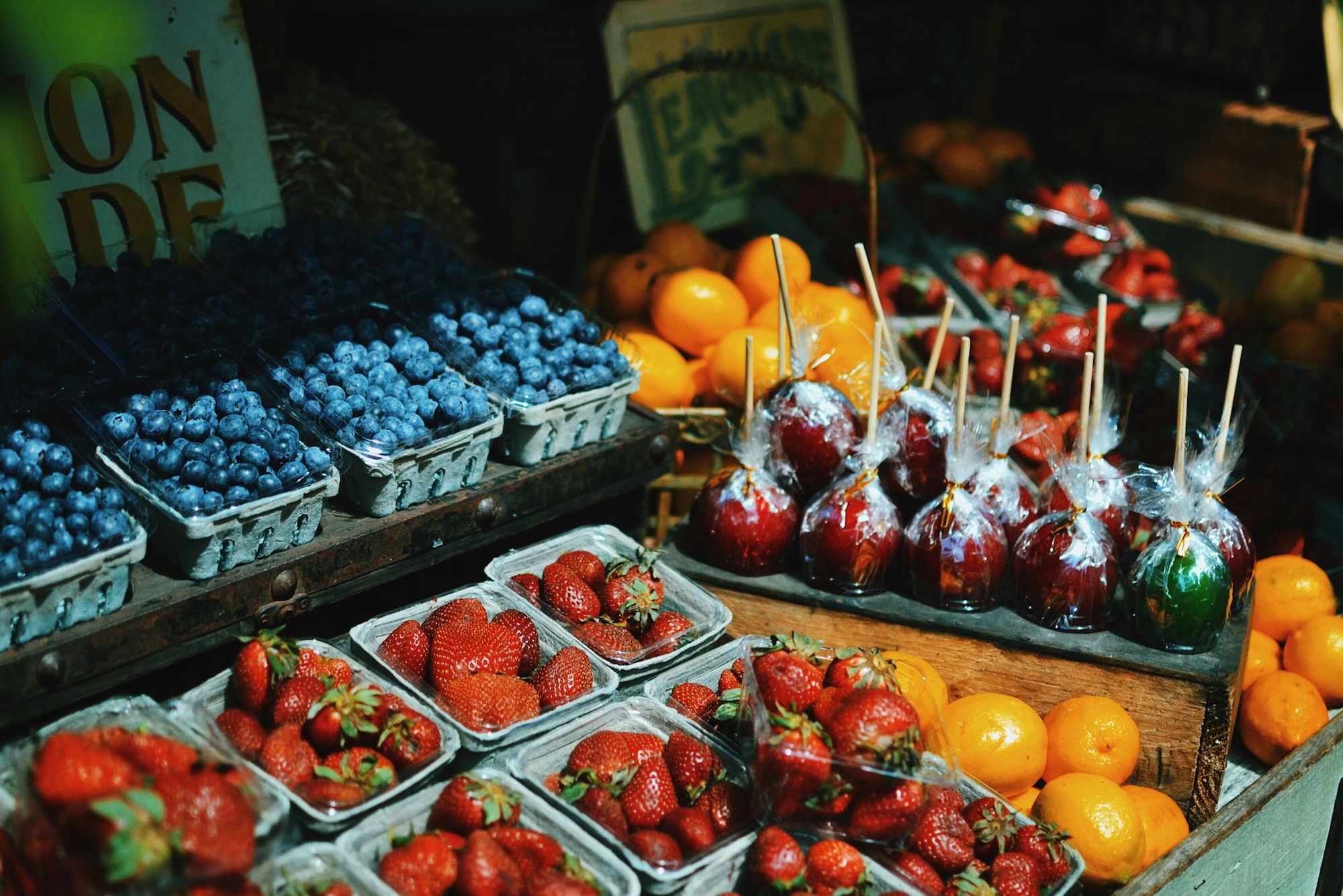 A market stall displaying blueberries, strawberries, apples on sticks, and oranges in baskets with a rustic wooden background.