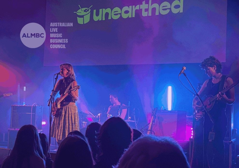 Band performing on stage with colorful lights, featuring a singer, drummer, and guitarist. "Unearthed" and "ALMBC" logos visible above.