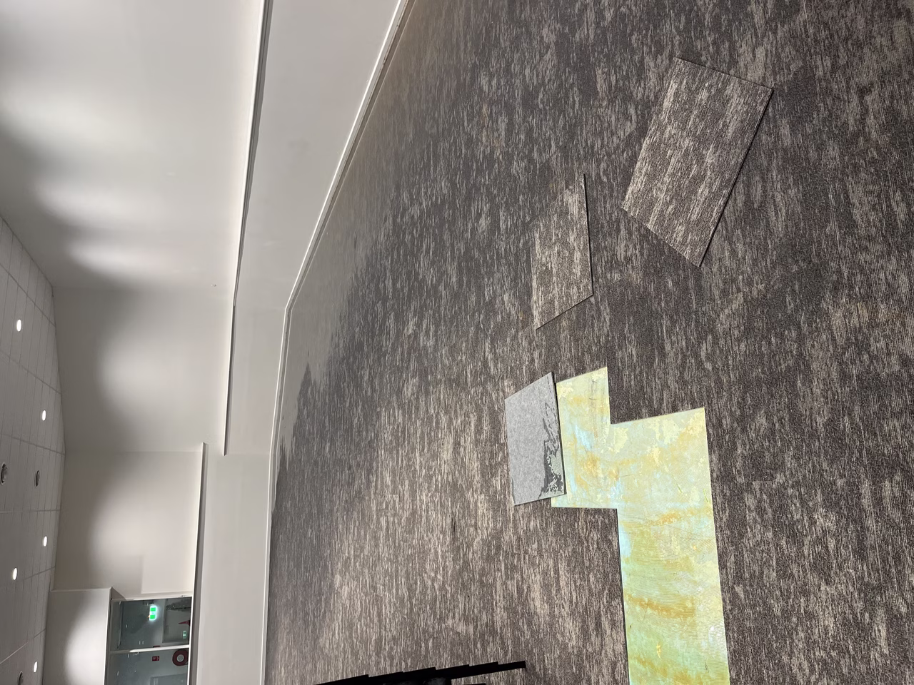 A room with gray carpet tiles, some lifted to reveal a colorful layer underneath, and white walls with a ceiling light. Flooded around the edges