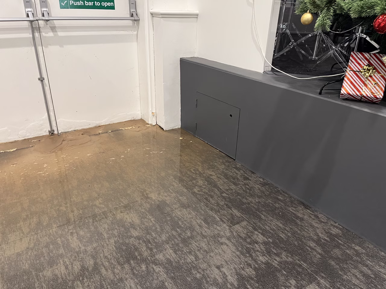 Water leaking onto a brown and gray floor near a gray wall with a Christmas tree and gift in the background.