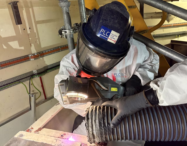 Precision Laser Cleaning Worker in protective gear and helmet using a tool to handle a flexible duct in an industrial setting, surrounded by scaffolding and equipment.