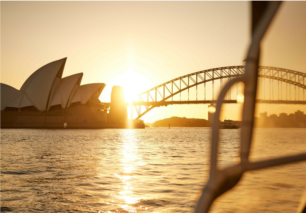 Sunset view of Sydney Opera House and Harbour Bridge from a boat, with warm golden light reflecting on the water.