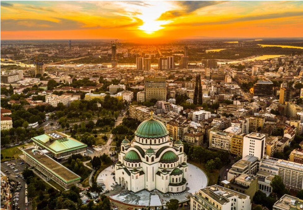 Aerial view of Belgrade city at sunset, featuring a large domed cathedral surrounded by buildings and greenery, with a river in the background.