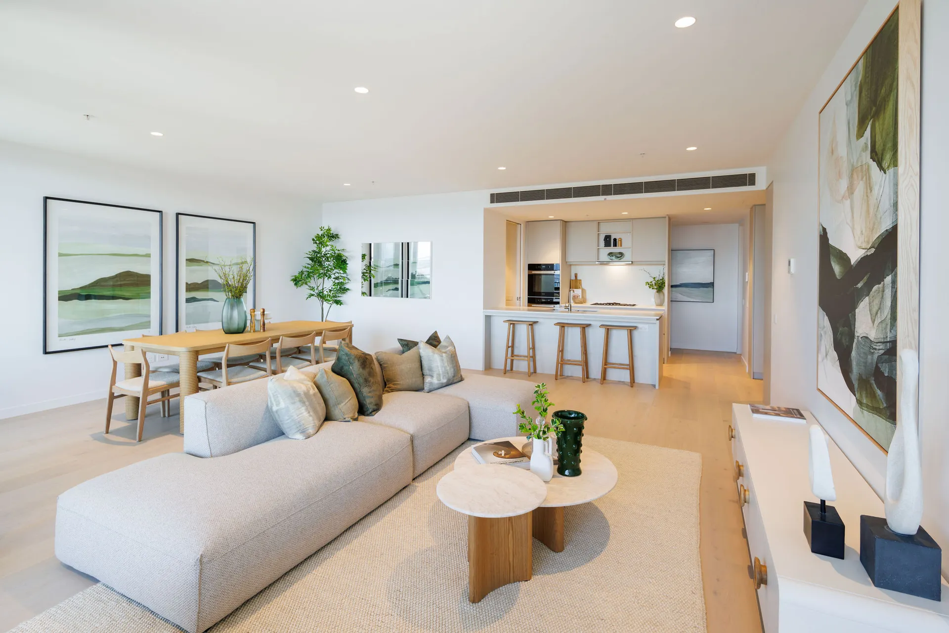 A spacious and contemporary open-plan living room seamlessly connects to a modern kitchen. The room features neutral tones with stylish decor, including a plush grey sofa, sleek wooden coffee table, and elegant light fixtures. Large windows allow natural light to flood the space, highlighting the minimalistic yet cosy ambiance.