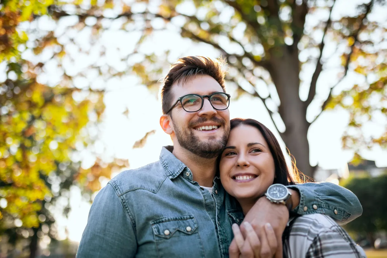 A couple smiling warmly as they embrace under a tree, with dappled sunlight filtering through the leaves casting gentle patterns of light and shadow around them.