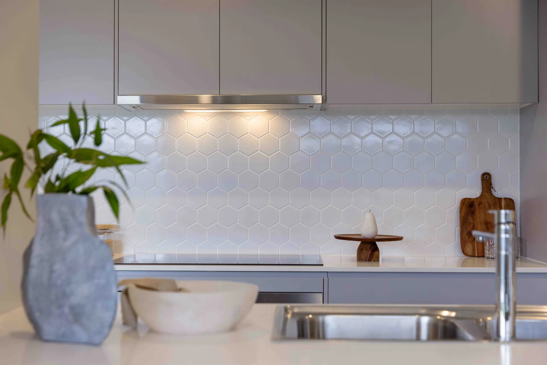 Modern kitchen with hexagonal white tile backsplash, grey cabinets, and a stainless steel sink. Décor includes a plant, vase, and wooden cutting boards.