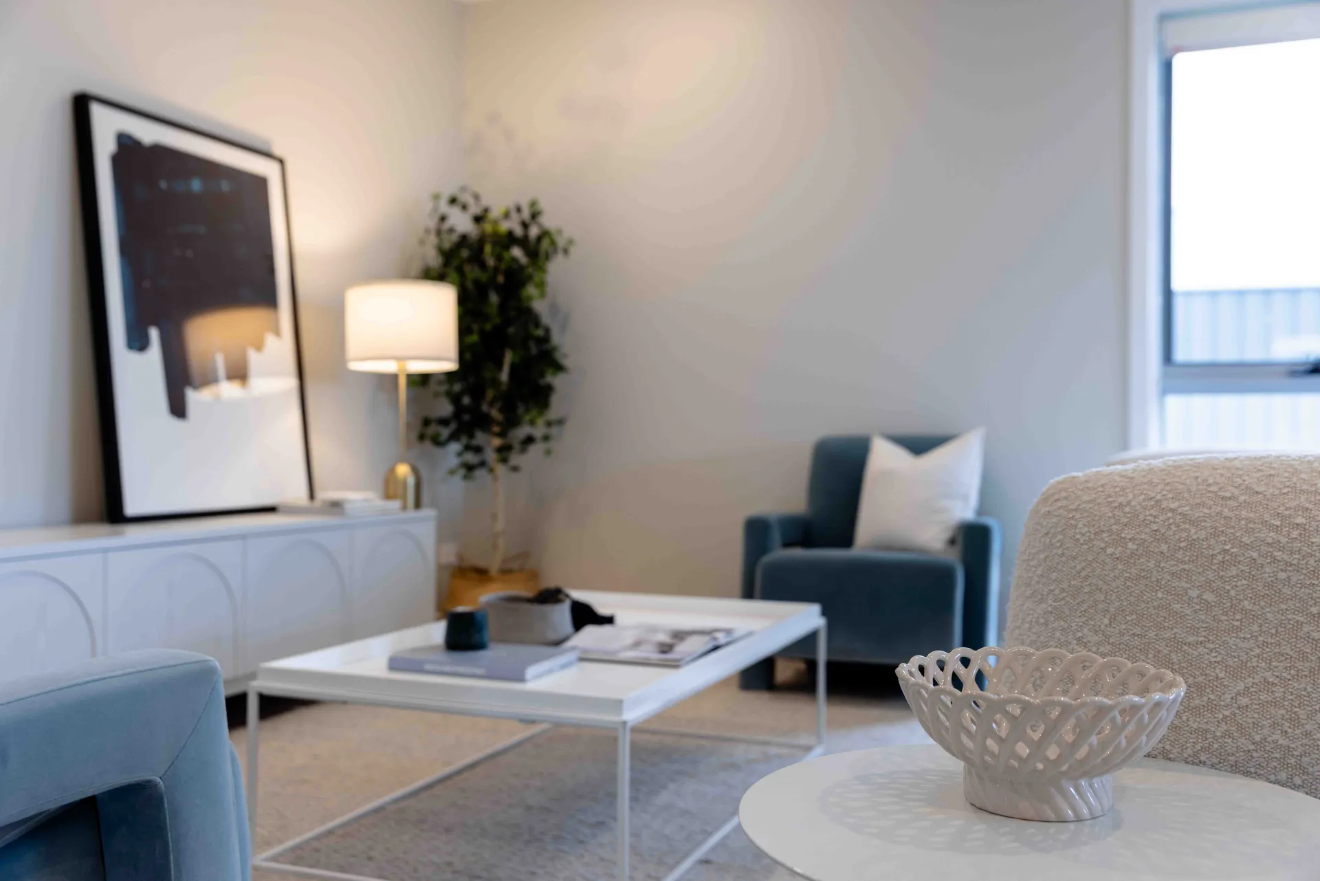 Modern living room with a blue armchair, white coffee table, abstract art, and a potted plant. Soft lighting and neutral tones create a cosy ambiance.