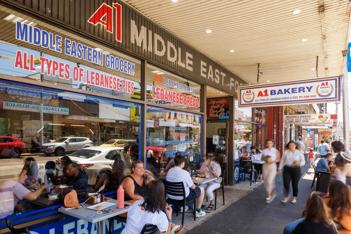 A bustling scene outside a Middle Eastern bakery, where several people are seated at small tables on the footpath, enjoying their meals. The atmosphere is lively, with a mix of locals and tourists savouring aromatic dishes. The street is lined with shops and cafes, creating a vibrant urban backdrop under a clear blue sky.