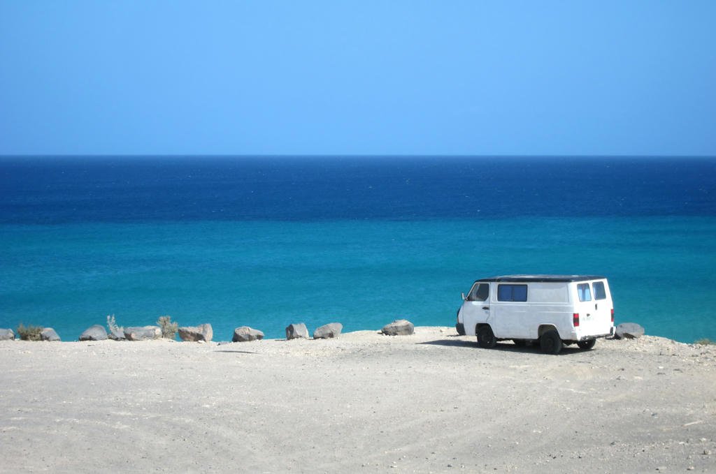 A white van parked on a sandy beach, overlooking a calm blue ocean under a clear sky.