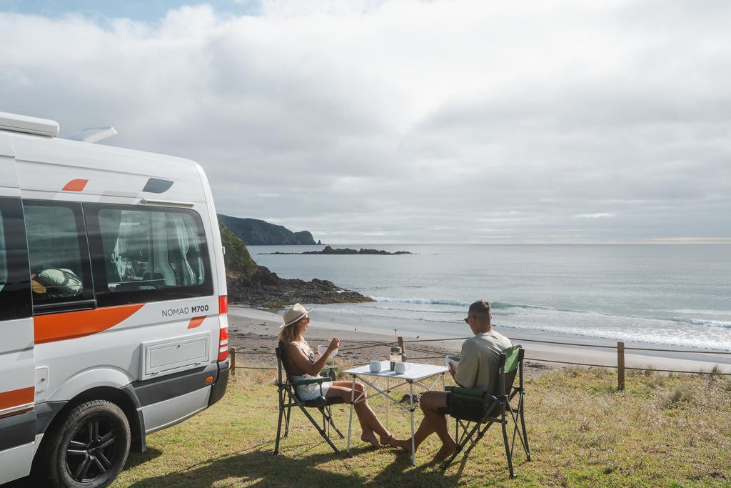 A couple sits by a camper van at a coastal campsite, enjoying drinks at a table with a scenic ocean view under a cloudy sky.
