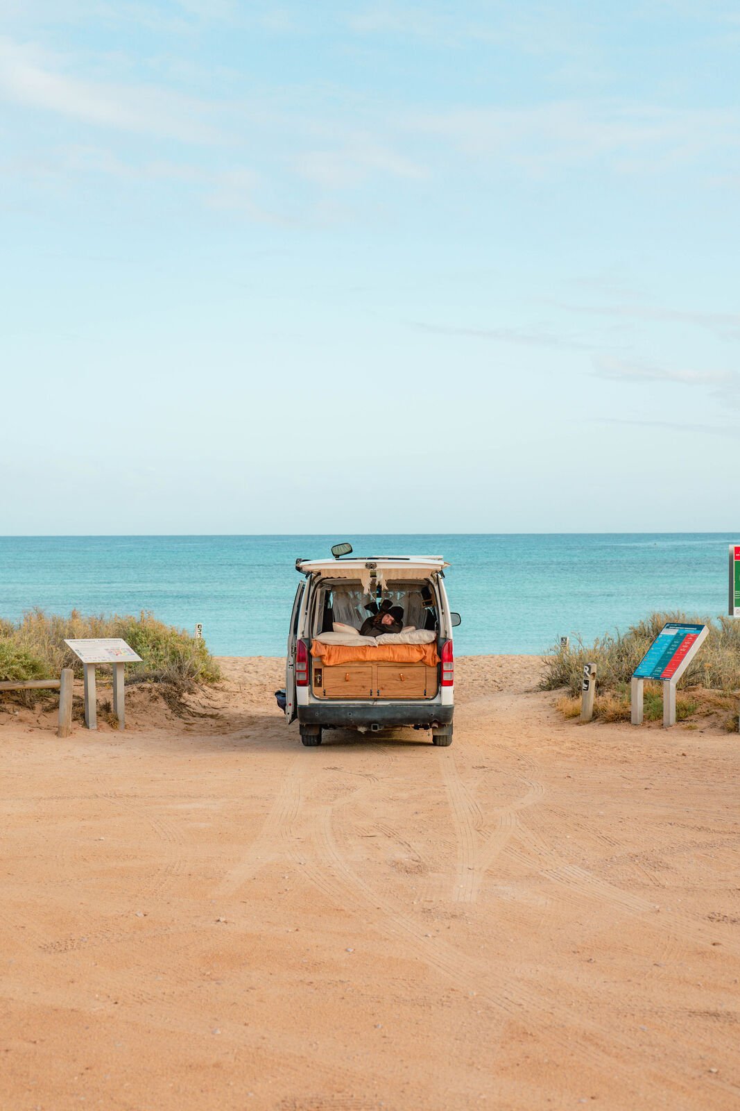 A camper van parked on a sandy beach with ocean views. The back is open, revealing a person relaxing on a bed inside.