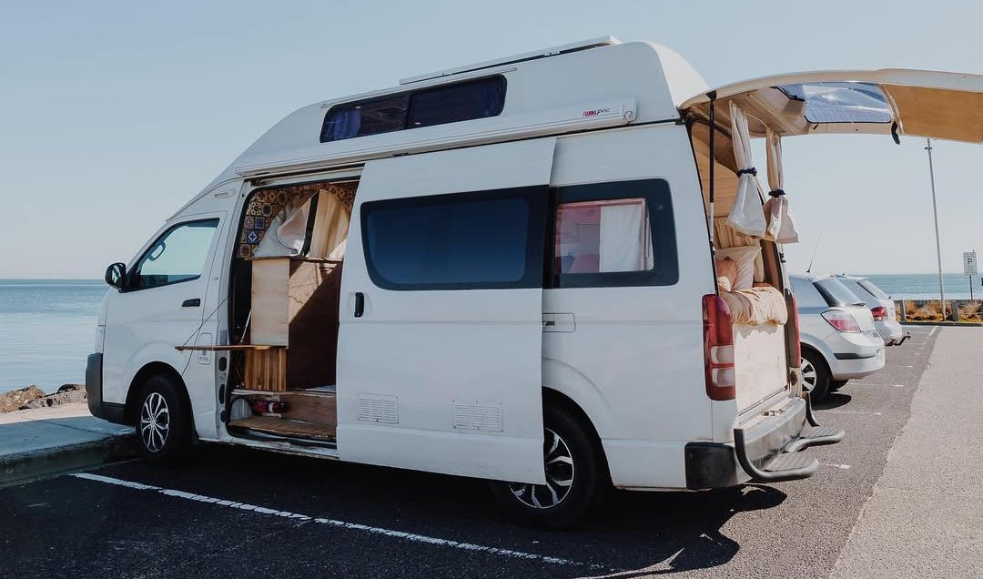 White camper van parked by the sea, with side and rear doors open, revealing a cozy interior. Clear blue sky and ocean in the background.