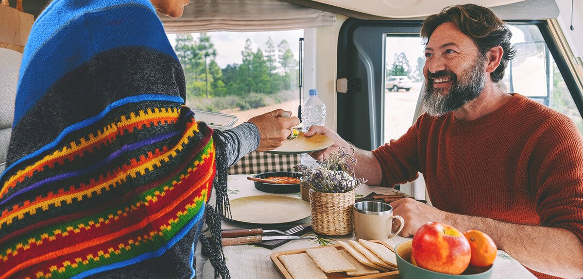 Autocaravana estacionada; pareja cocinando al aire libre con montañas al fondo. Comida y viaje.