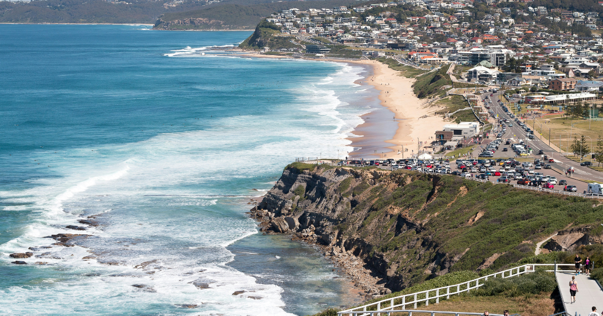 Coastal view of a busy road alongside a cliff, with waves crashing on the shore and a town visible in the background.