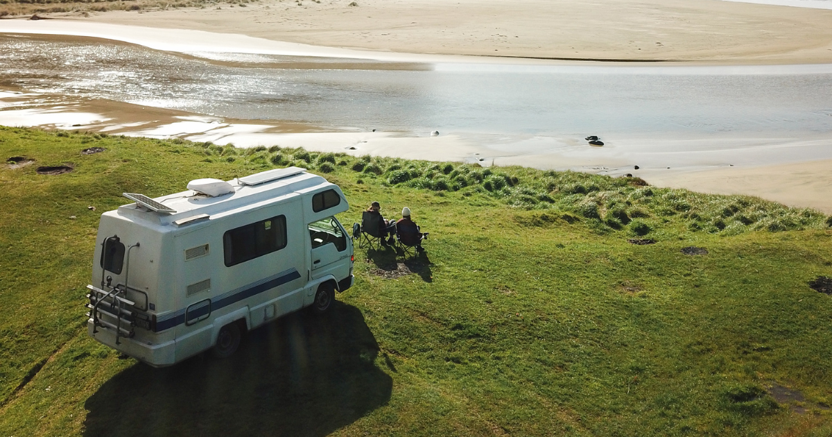 A camper van parked on a grassy hill by a river, with two people sitting in chairs enjoying the view of the sandy beach and water.