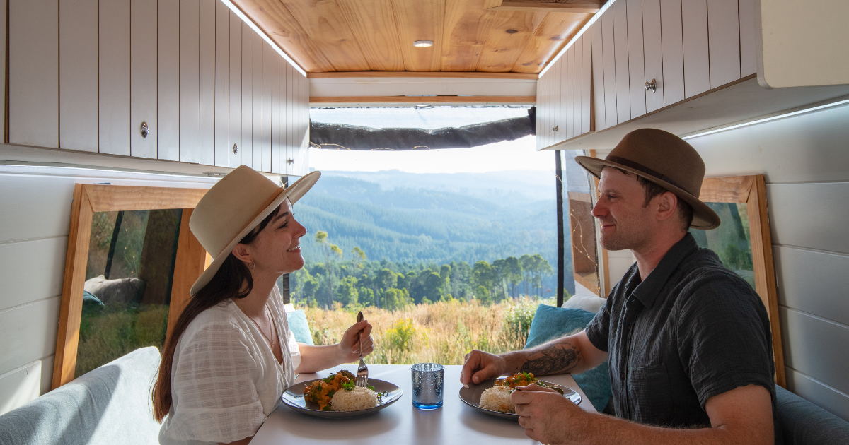A man and a woman sitting in a campervan at a table, eating dinner. The campervan doors are open and you can see mountains in the background