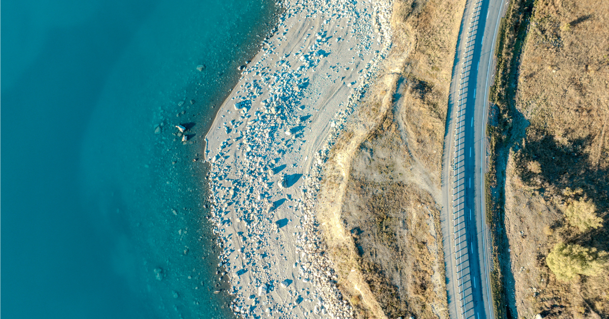 Aerial view of a winding road beside a rocky shoreline with clear blue water and dry, grassy terrain.