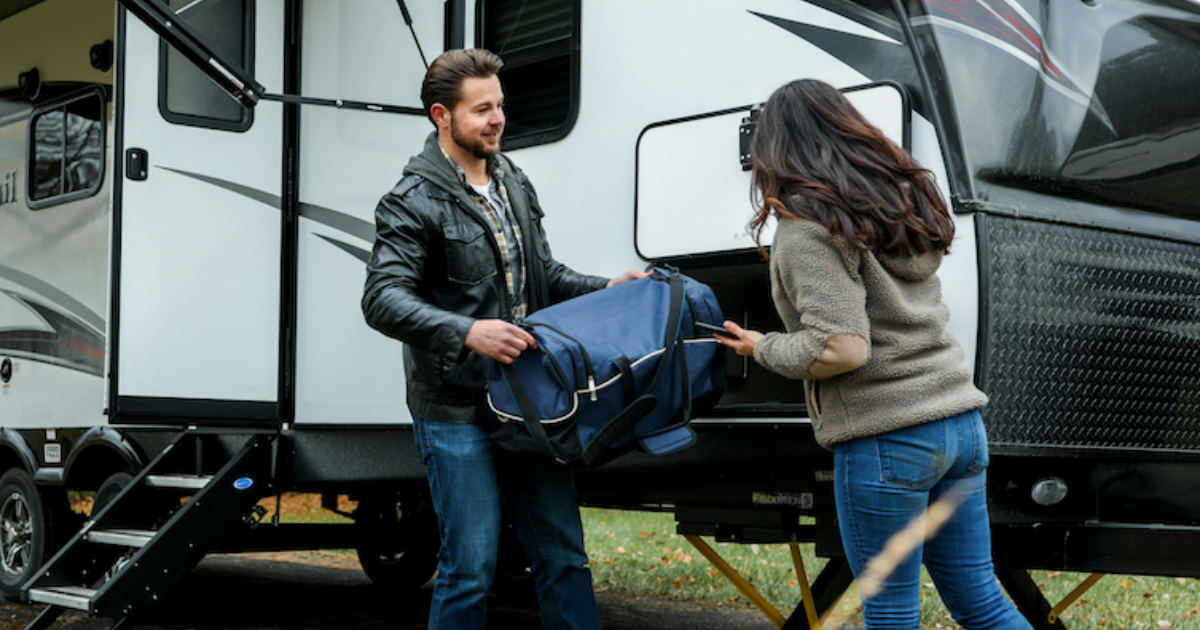 A man and woman unload a blue bag from a camper trailer, with steps visible on the left and the vehicle's exterior details in view.