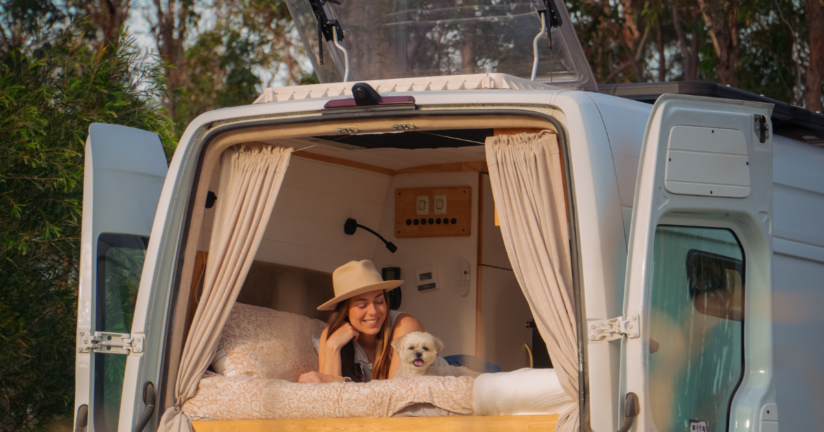 Woman in a hat and a small dog relax in a camper van with open doors, surrounded by trees.