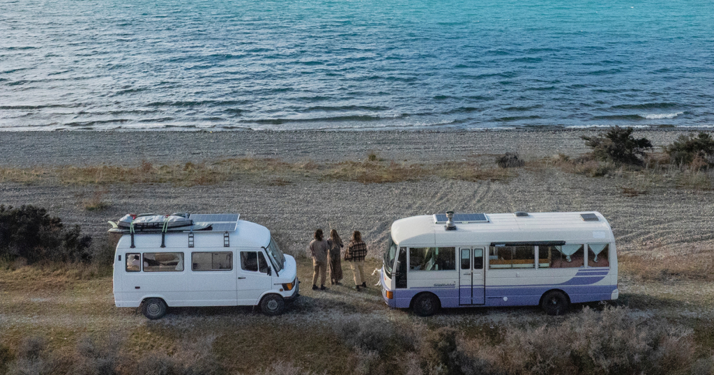 Two vans parked on a gravel beach near the sea, with three people standing between them, facing the water.