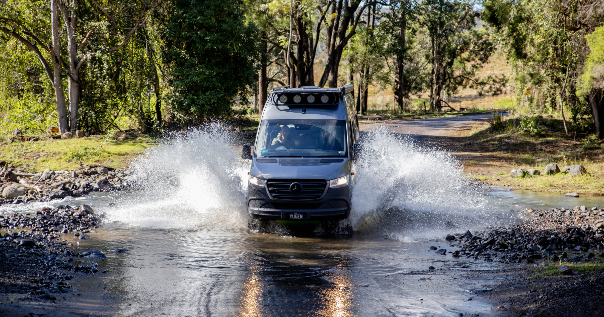 A van drives through a shallow stream on a forest road, creating splashes of water. Trees and greenery surround the scene.