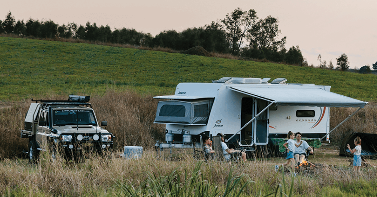 A family relaxes by a campfire near a white caravan and parked SUV in a grassy field, with trees in the background.