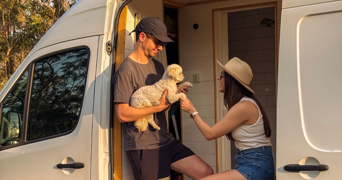 A man and woman with a small white dog sit in the doorway of a white van, enjoying a sunny day outdoors.