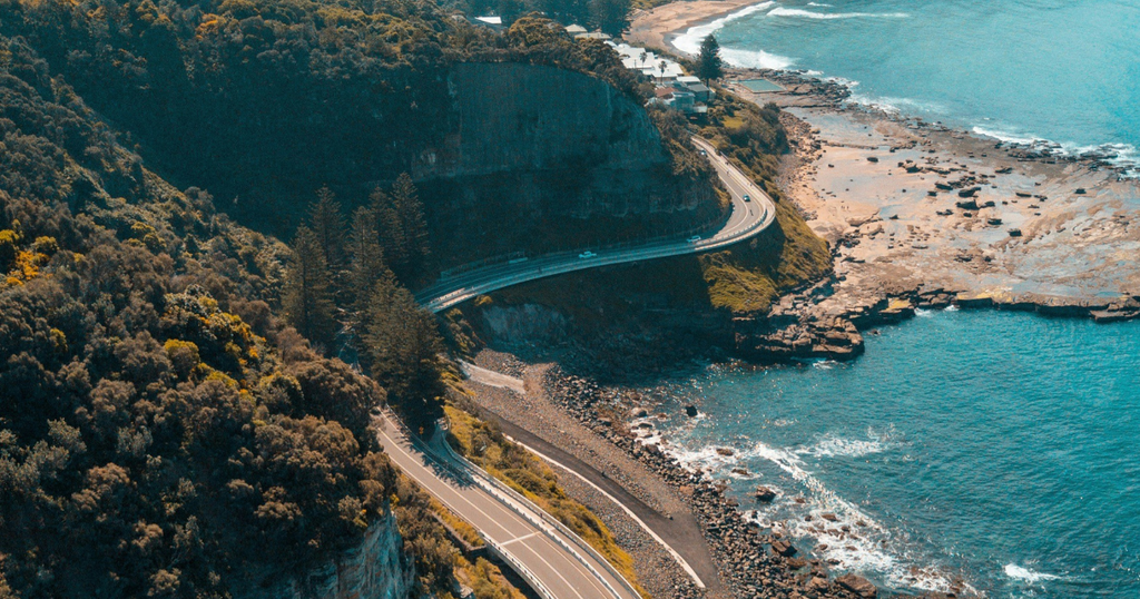 Aerial view of a coastal road winding along a cliff, surrounded by lush greenery and clear blue ocean waters.