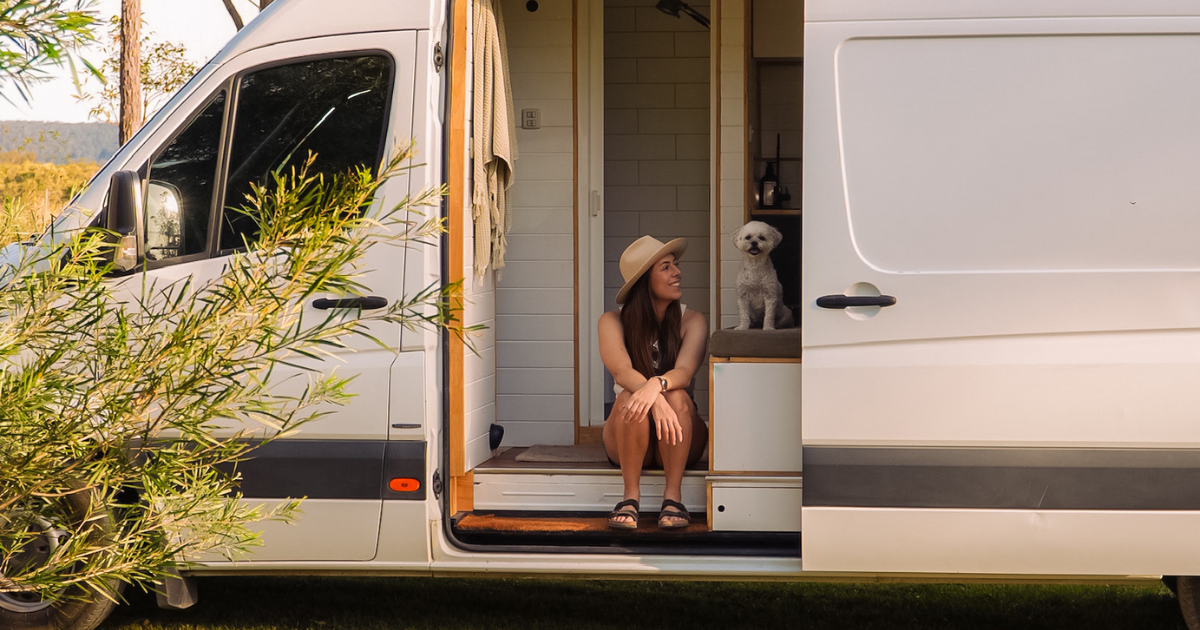 Woman sitting in a camper van doorway with a small white dog beside her, surrounded by greenery and sunlight.