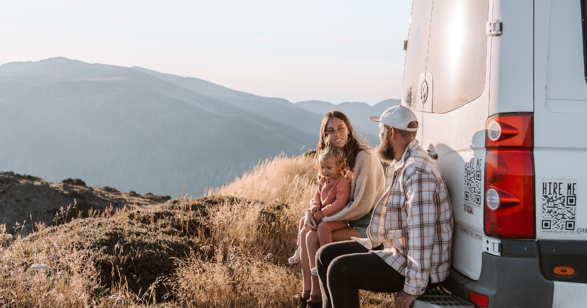 A family of 3 people sitting on the back of a campervan overlooking the mountains
