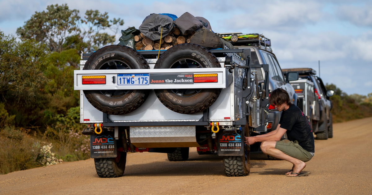 A person secures a trailer loaded with spare tires and supplies on a dirt road, with two off-road vehicles in the background.