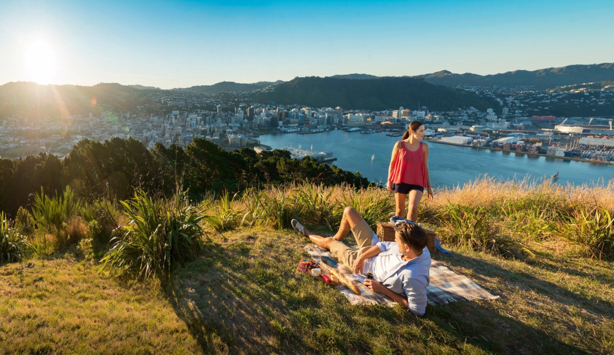 A couple enjoys a picnic on a grassy hill overlooking a cityscape and harbor at sunset, with mountains in the background.