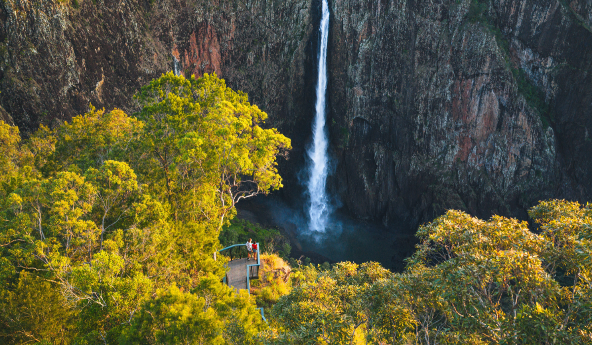 Sunlit trees frame a tall narrow waterfall plunging into a rocky gorge while two people stand on a viewing platform.