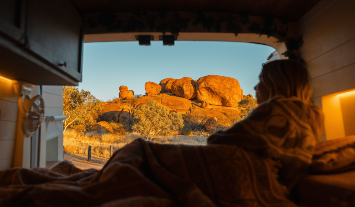 Person wrapped in a blanket inside a camper van, looking out at sunlit red rock formations.