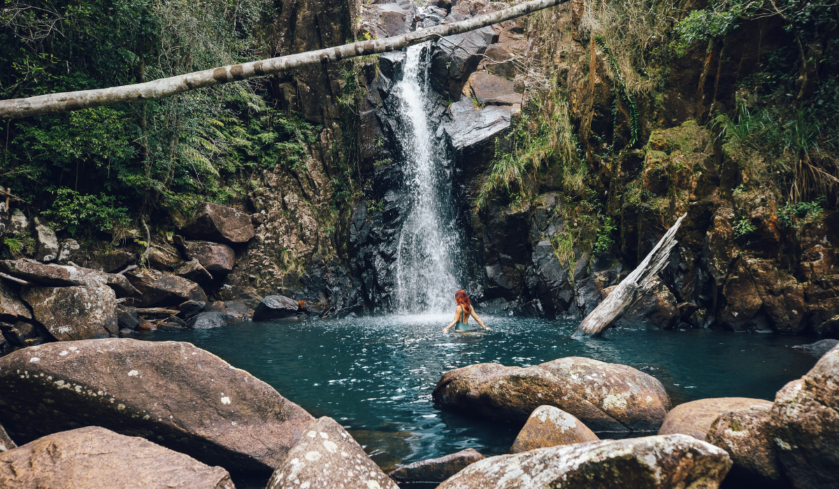 Person with red hair wading in a turquoise pool beneath a waterfall in a rocky forest gorge.