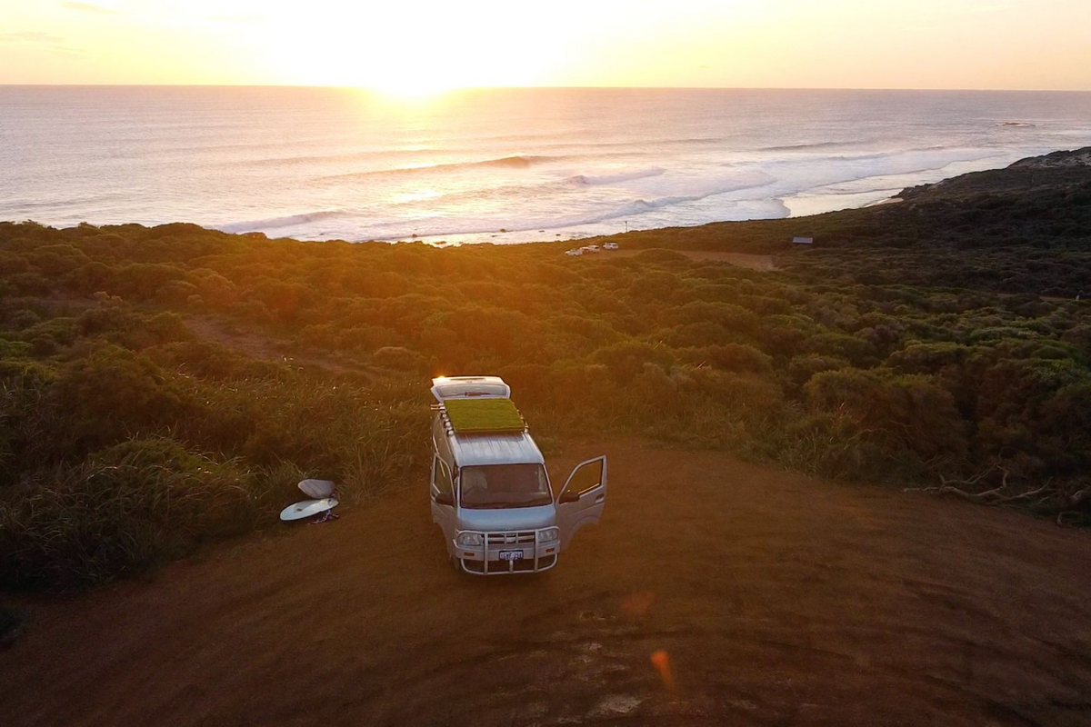A van parked on a coastal dirt path with surfboards nearby, overlooking the ocean at sunset. Lush greenery surrounds the area.