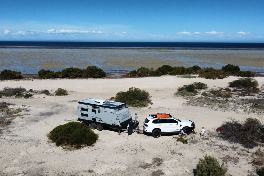 A white SUV towing a camper is parked on a sandy coastal area with sparse vegetation, under a clear blue sky.