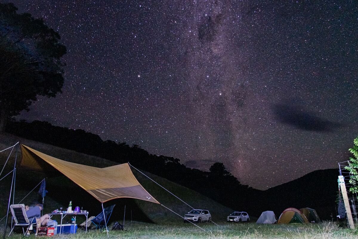 Starry night sky over a campsite with tents, a tarp, and a parked car in a grassy area surrounded by hills.