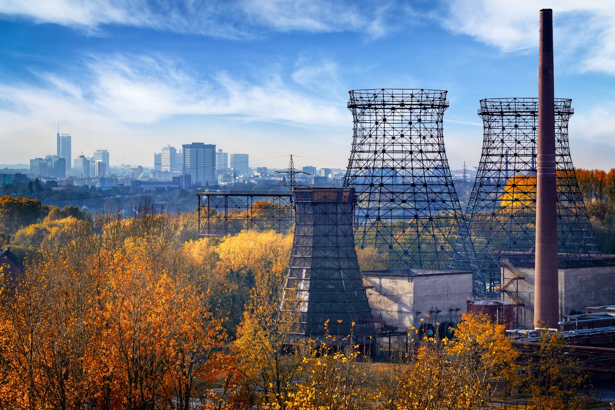 Industrielle Kühltürme und ein Schornstein ragen über herbstliche Bäume hinaus, mit einer Stadtsilhouette im Hintergrund unter einem blauen Himmel.