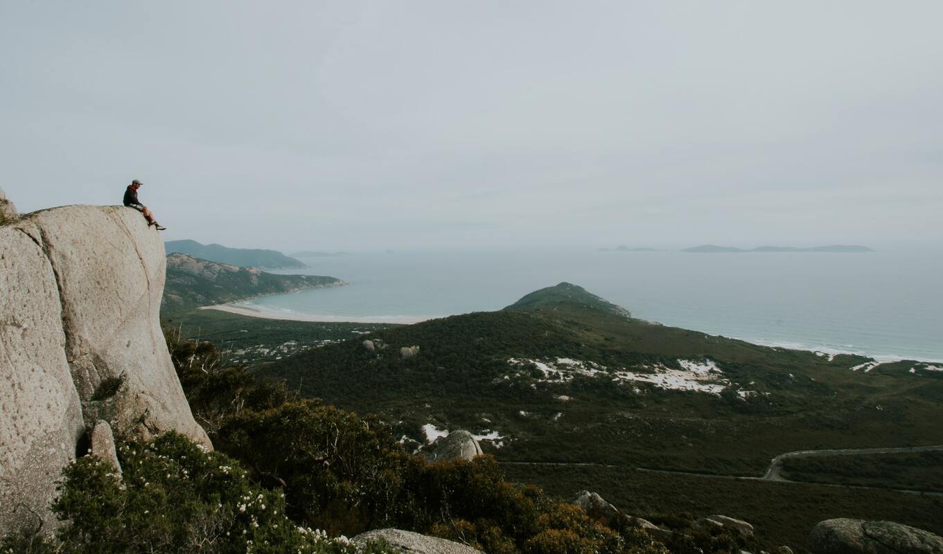 Person sitting on a cliff edge overlooking a vast coastal landscape with greenery, hills, and the ocean under a cloudy sky.
