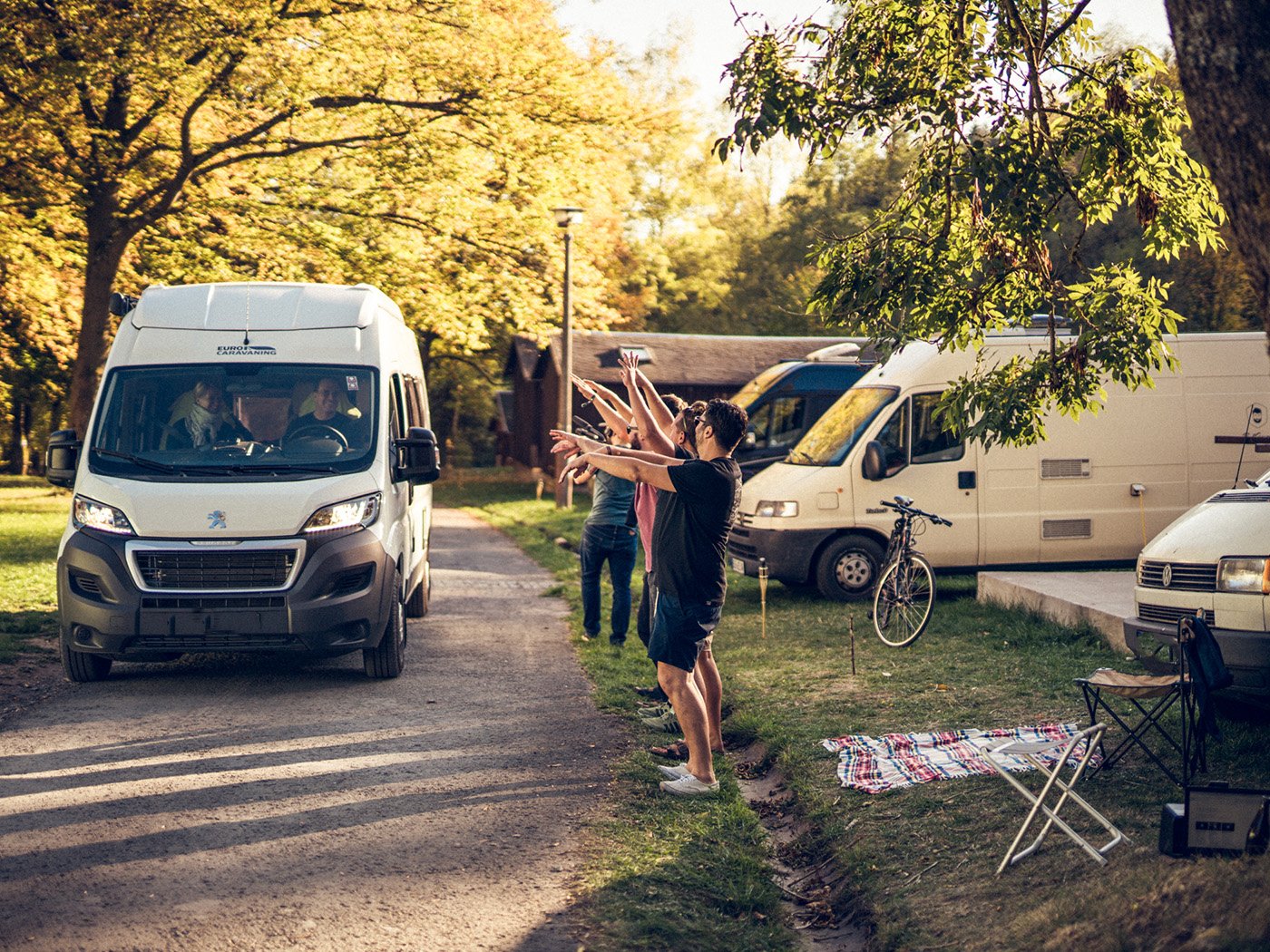 Ein weißer Camper fährt an anderen Campern auf einem Campingplatz vorbei. am Rand stehen Menschen und machen eine Laola