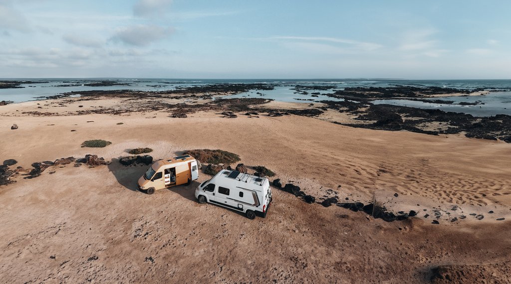 Dos furgos aparcadas en una playa de arena cerca de costas rocosas, con un vasto océano y cielo nublado de fondo.