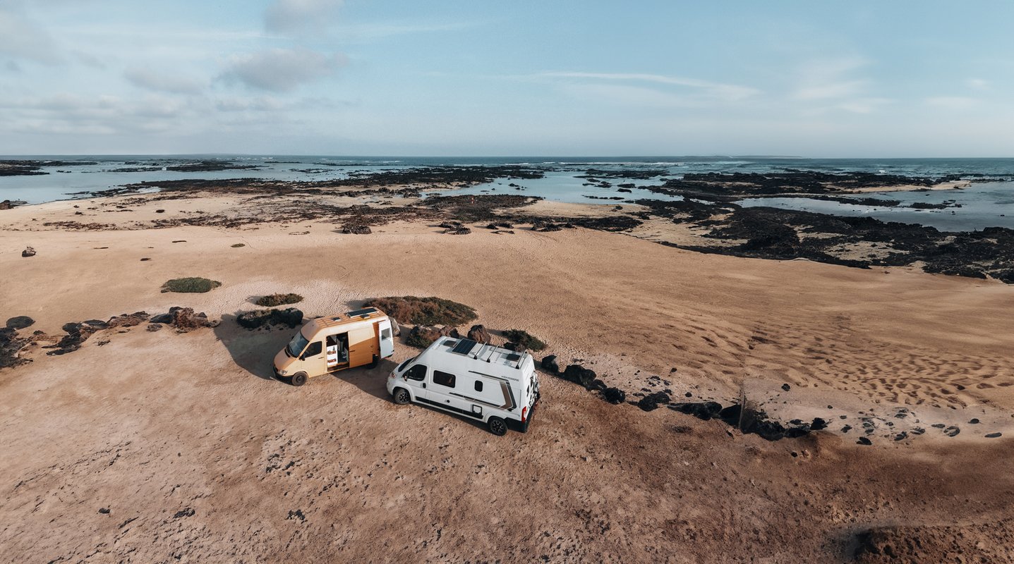Dos furgos aparcadas en una playa de arena cerca de costas rocosas, con un vasto océano y cielo nublado de fondo.