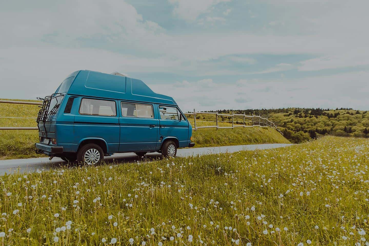Blauer Campervan, der auf einer Landstraße geparkt ist, umgeben von einem grünen Feld mit weißen Blumen unter einem bewölkten Himmel.