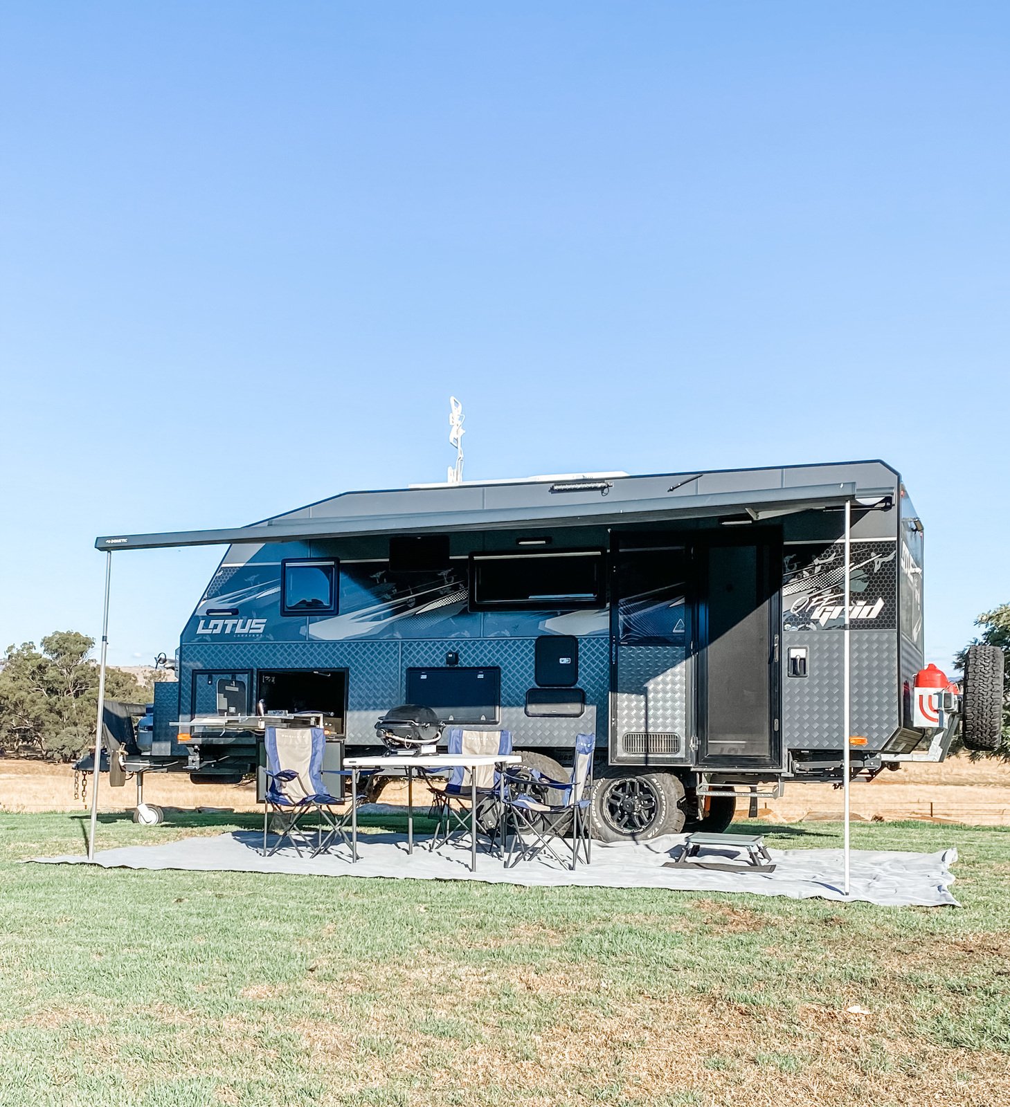Lotus Caravan hired on Camplify with awning, outdoor chairs, and table set up on grassy field under clear blue sky.