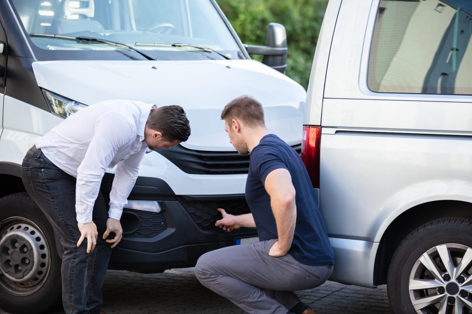 Two men inspecting the bumper area between a white van and a silver van, possibly assessing damage or discussing repairs.