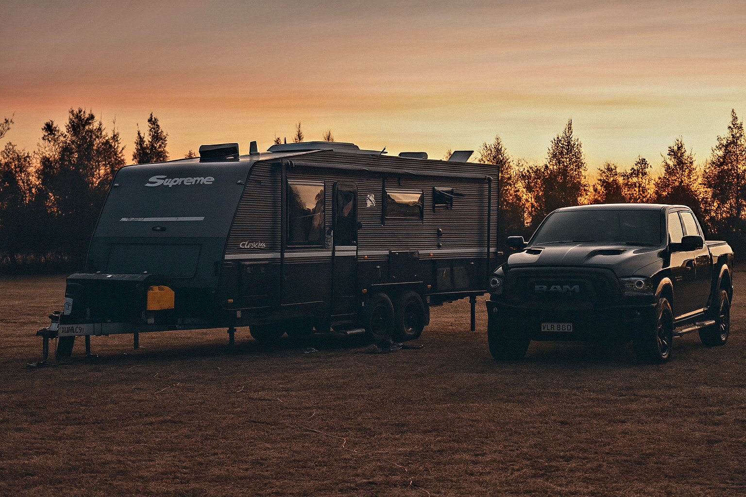 Black truck and Supremem caravan hired on Camplify parked on grass at sunset, surrounded by trees, under an orange sky.