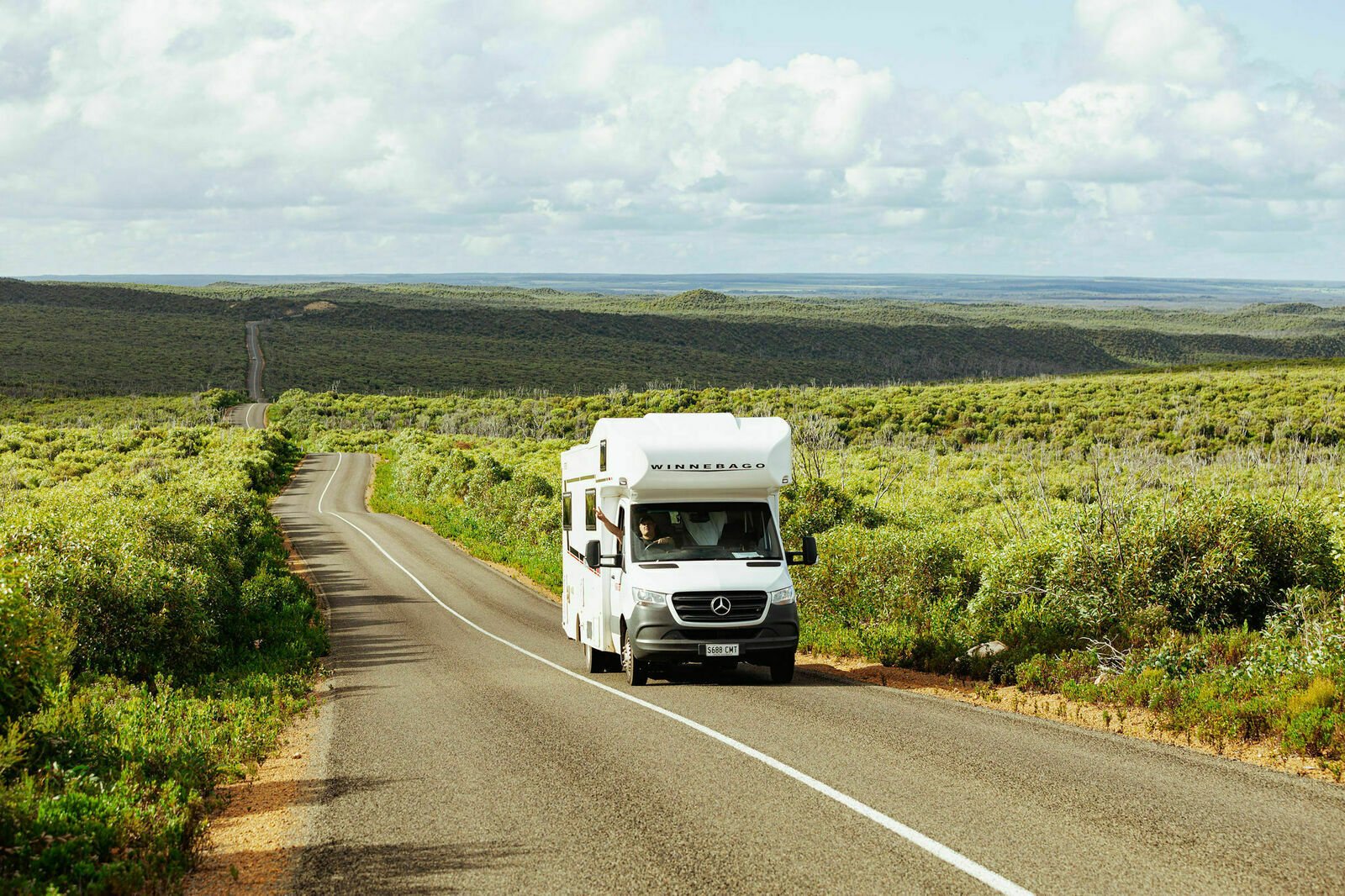 A white Winnebago motorhome hired on Camplify drives along a winding road through a lush, green landscape under a cloudy sky, surrounded by rolling hills.
