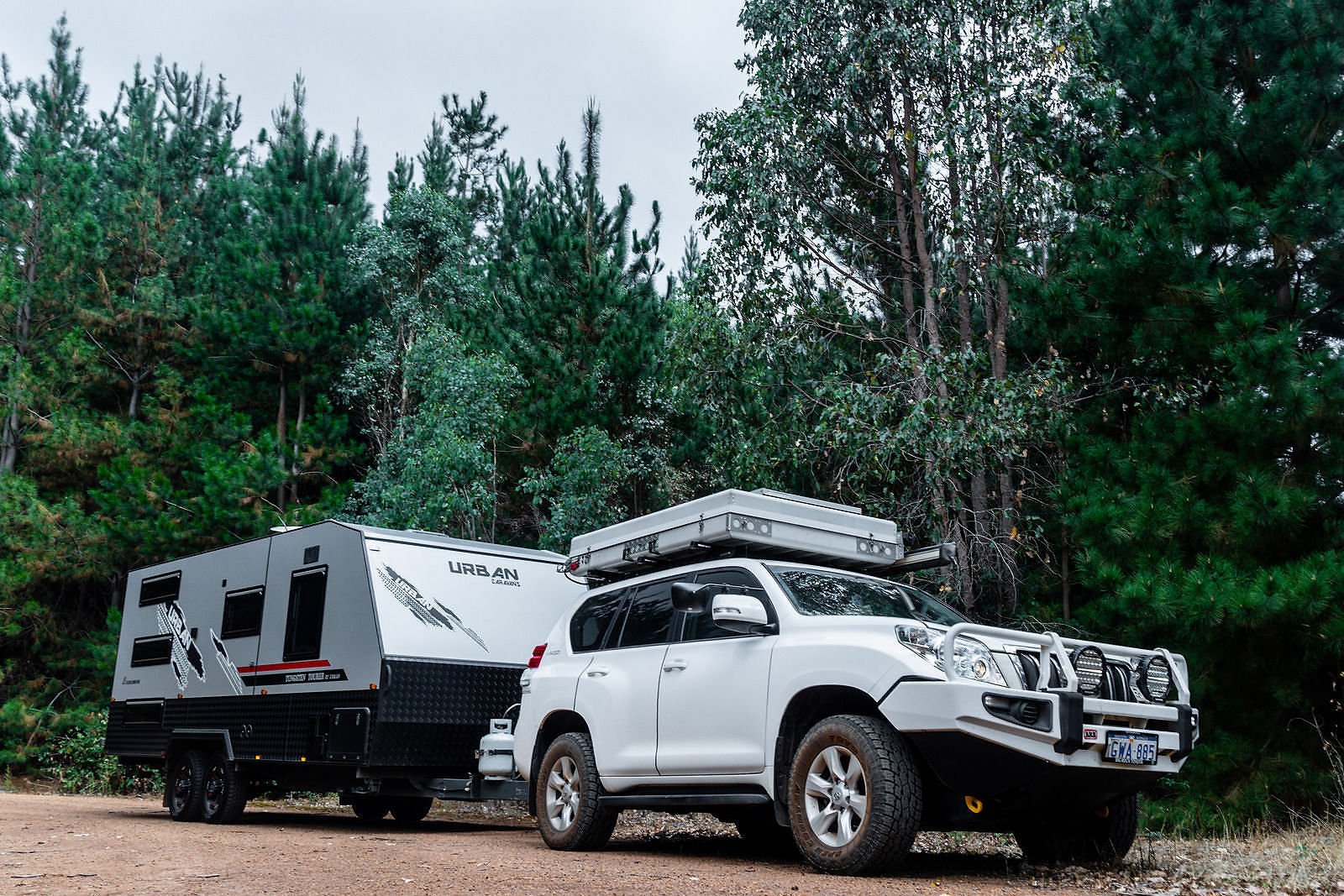 White SUV towing a gray caravan parked on a dirt road, surrounded by tall green pine trees under a cloudy sky.